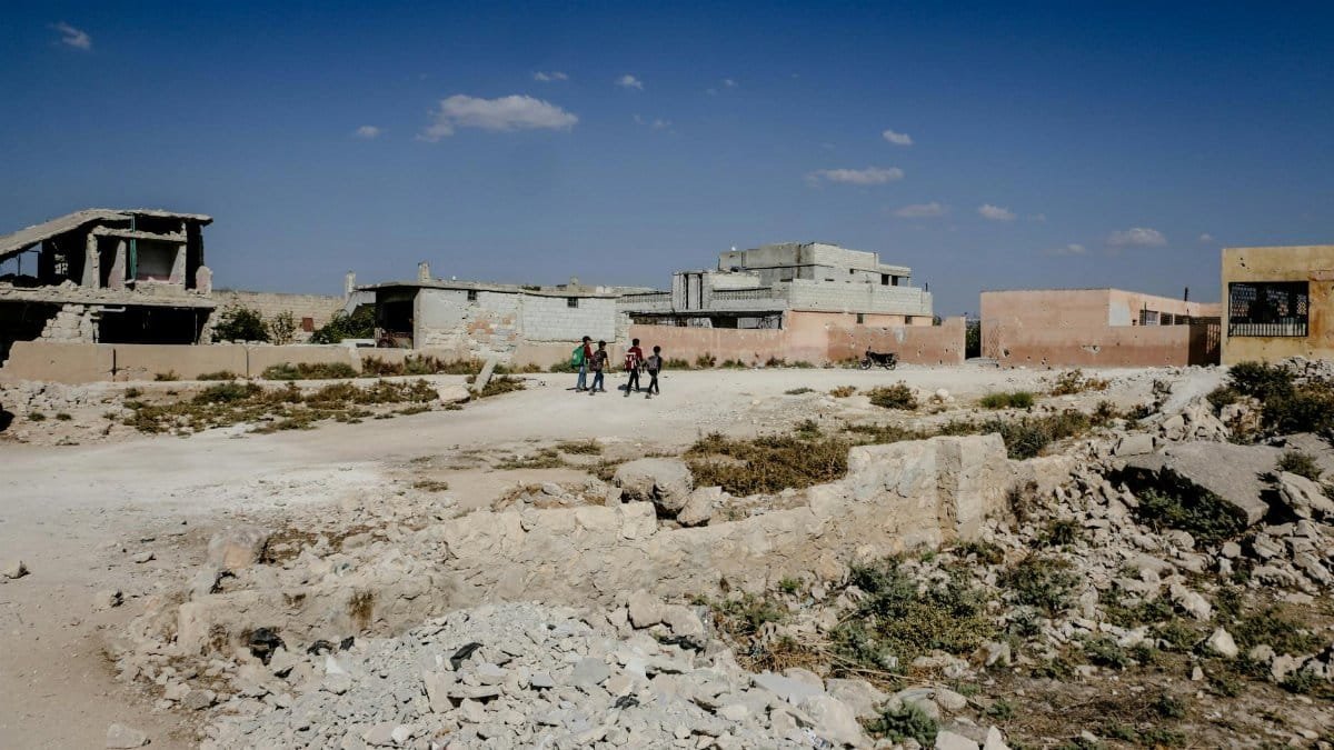 Children walking across a dirt pathway in war-torn Idlib, showcasing resilience amid destruction.