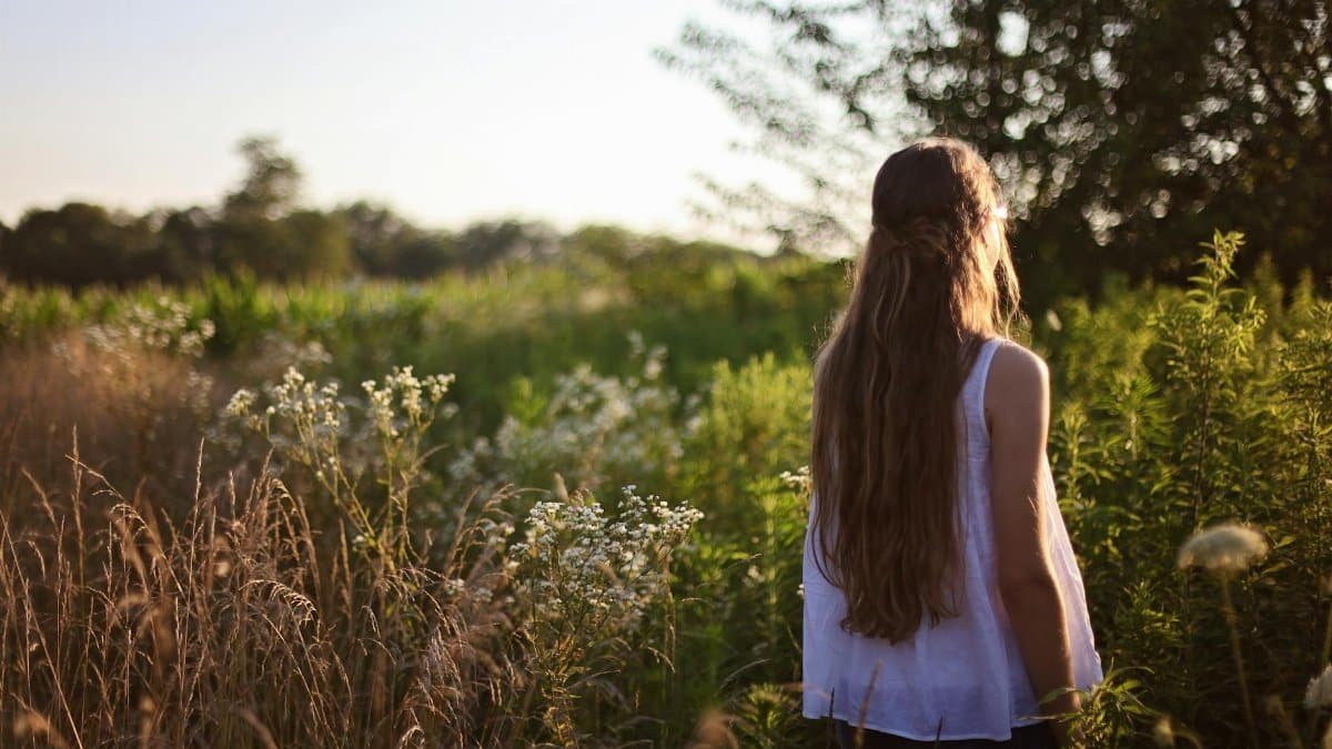 A woman with long hair stands in a sunlit countryside field surrounded by wildflowers.