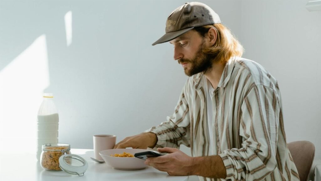 A man enjoys a casual breakfast with cereal in a sunlit room, using his phone.