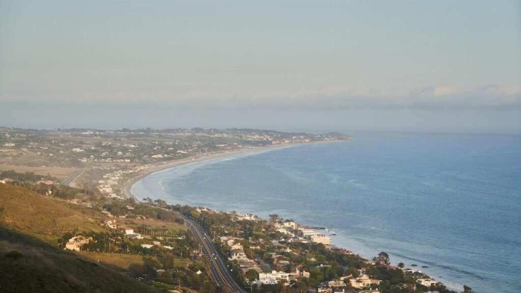 Scenic aerial view of a coastal highway running alongside a peaceful bay with distant hills and settlements.