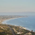 Scenic aerial view of a coastal highway running alongside a peaceful bay with distant hills and settlements.