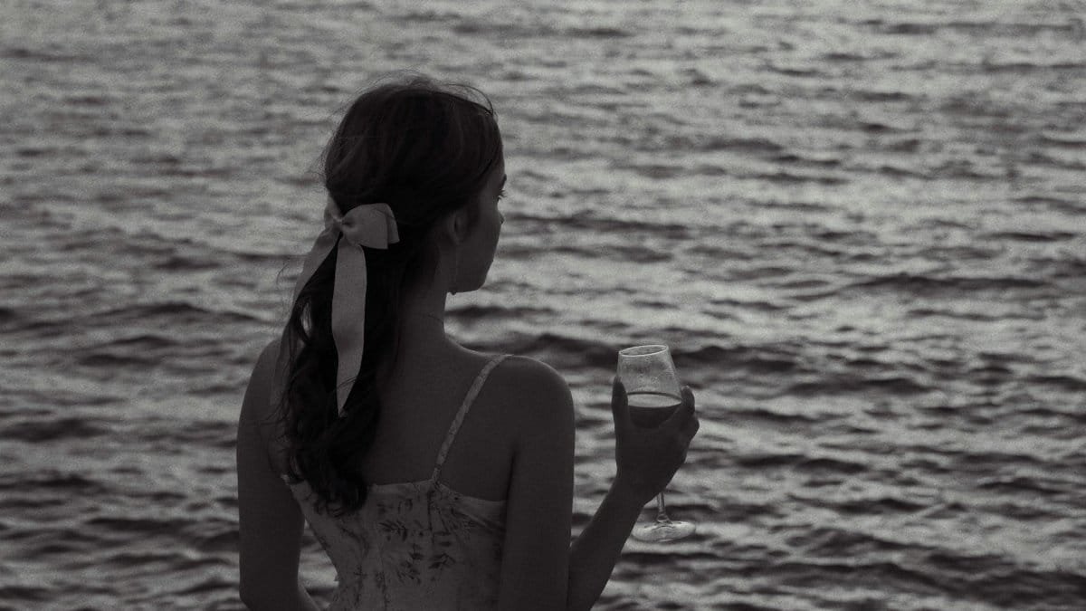 Young woman gazing at ocean with wine glass, serene and reflective mood.