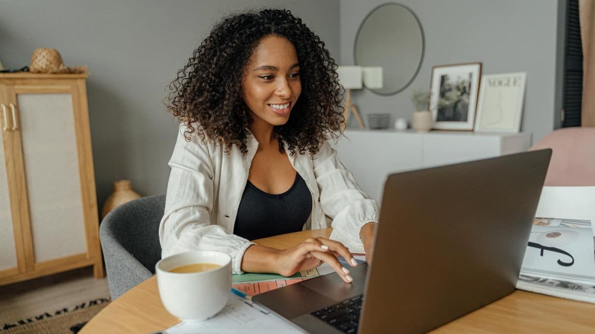 A woman sits at a round table, working on a laptop with a coffee cup nearby in a cozy home office setting.