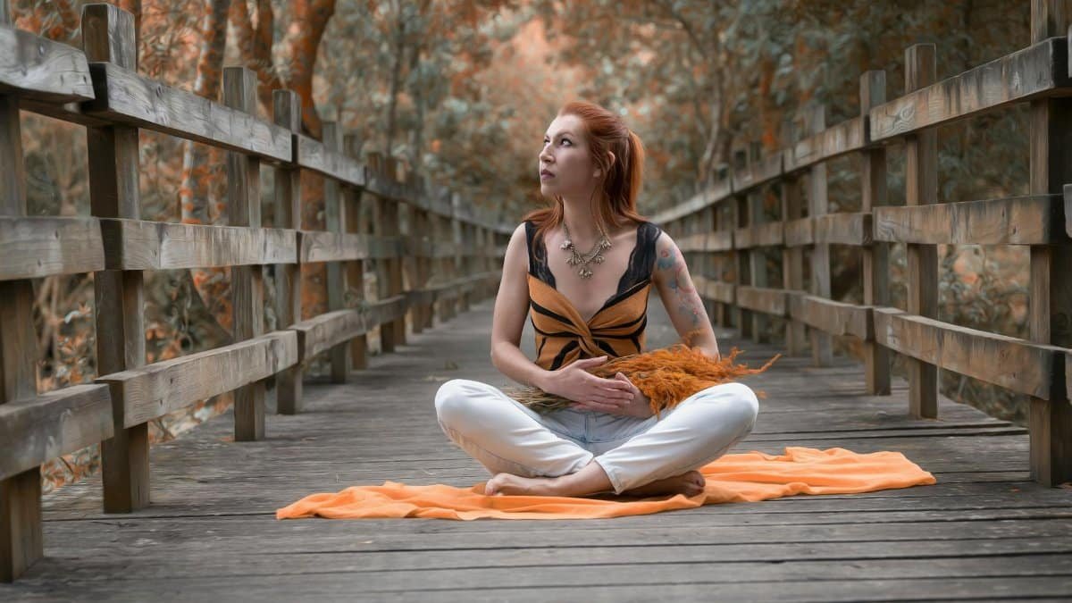 Redhead woman sits on a wooden bridge in nature, meditating under the trees.