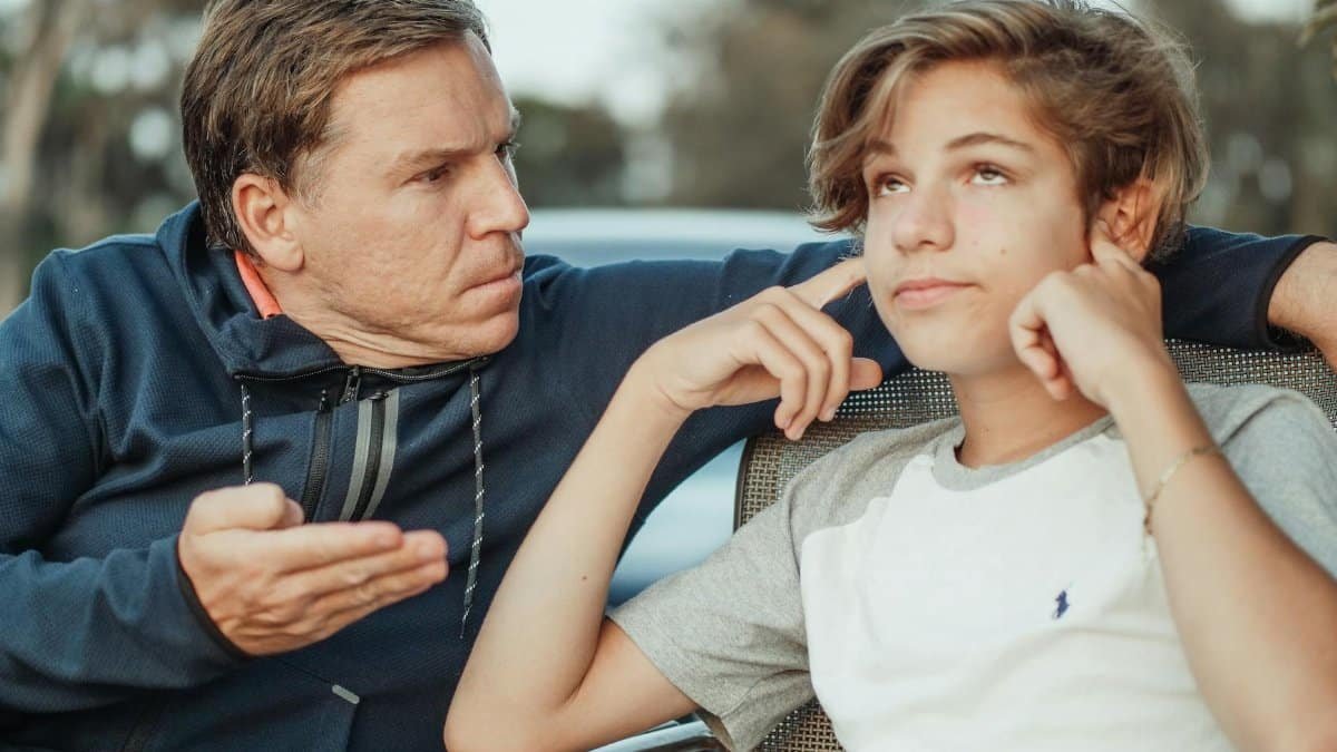 Father expressing frustration while teenage son shows defiance, seated outdoors.