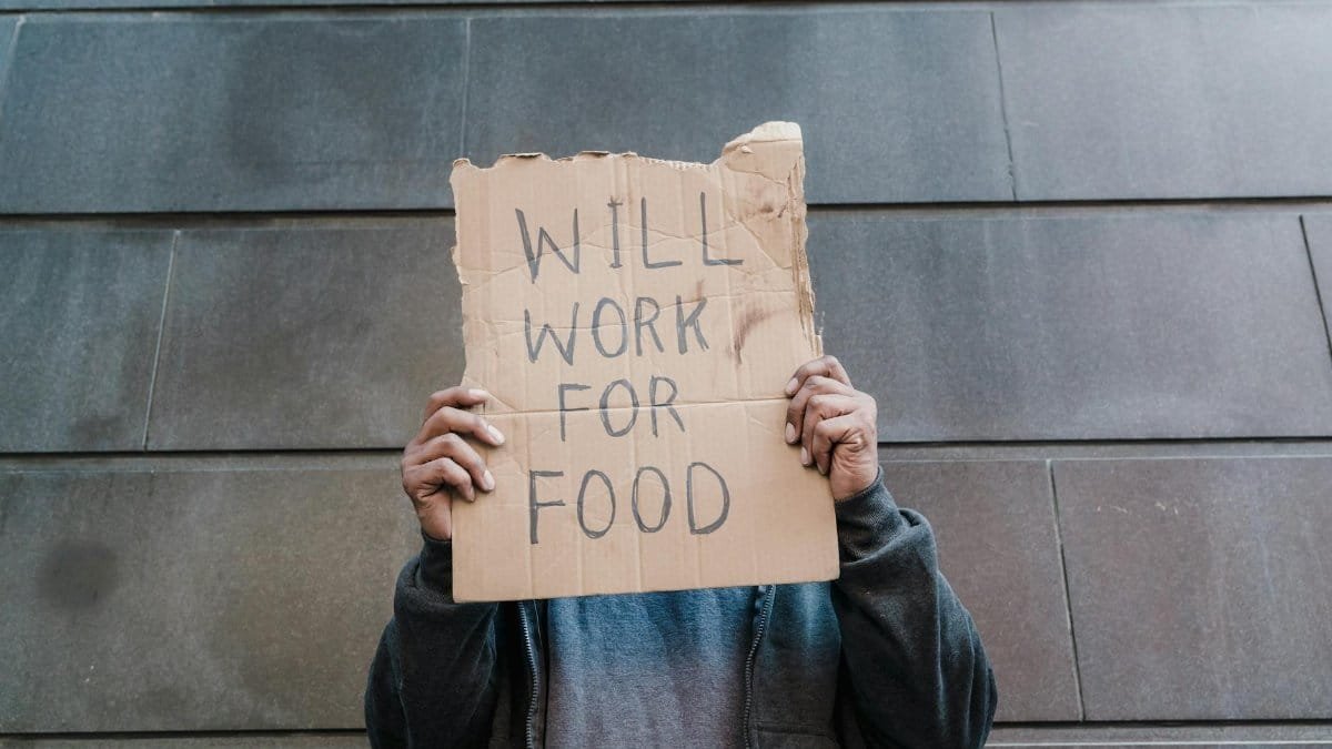 A person in a hoodie holding a 'Will Work for Food' cardboard sign against a wall.