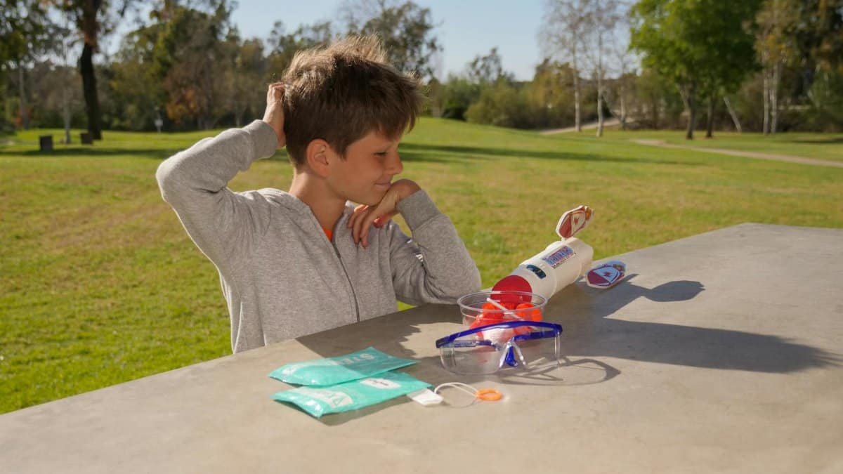 Boy in gray sweater plays with toy rocket at a park table on a sunny day.