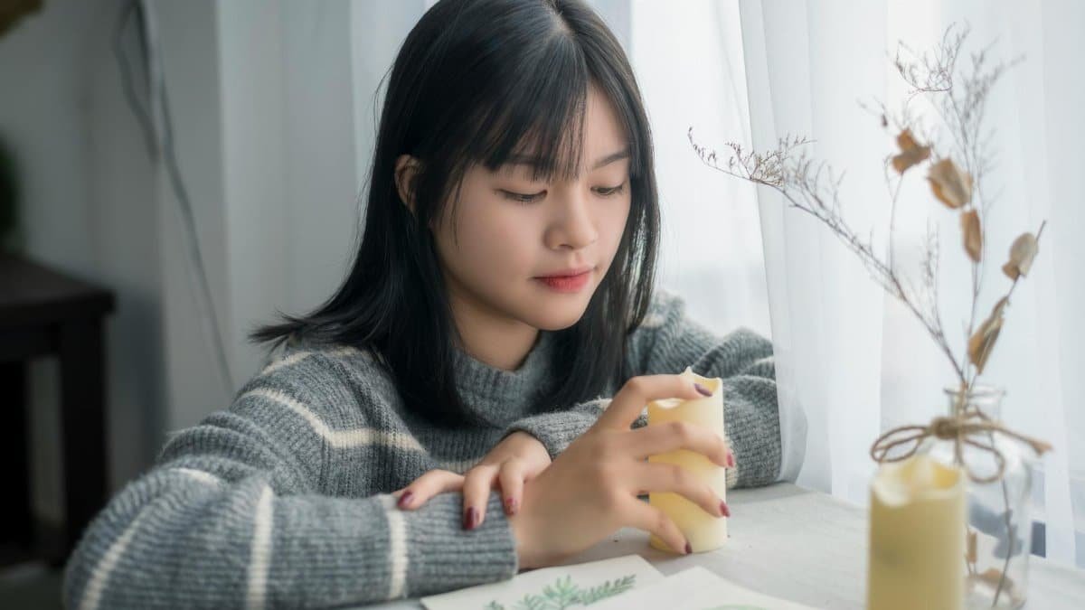 A young woman sits thoughtfully holding a candle in a cozy indoor setting.