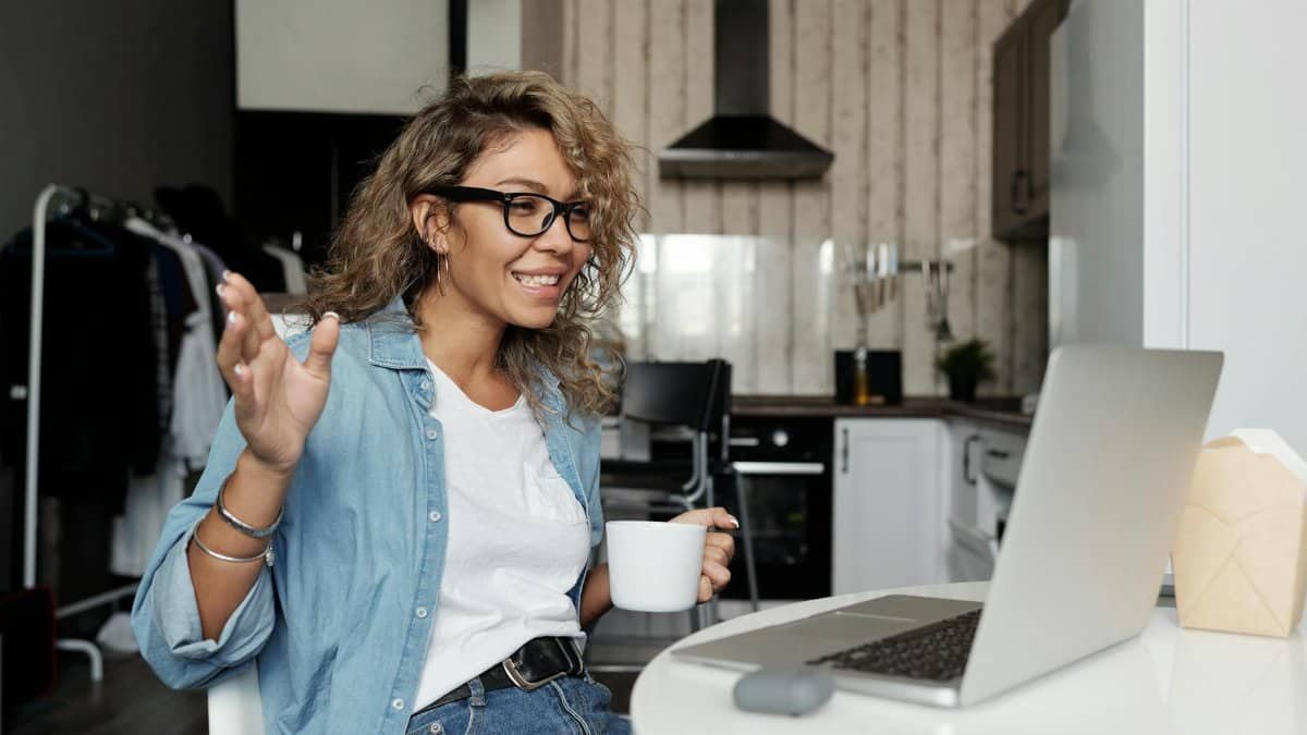 Smiling woman with glasses having a video call from home, enjoying a cup of coffee in her kitchen.