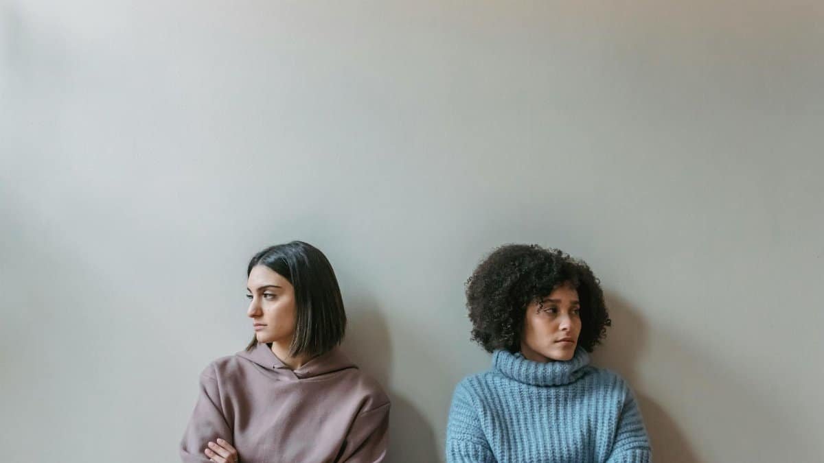 Sad multiracial female friends with crossed arms looking away and standing near white wall while having conflict in light room at home