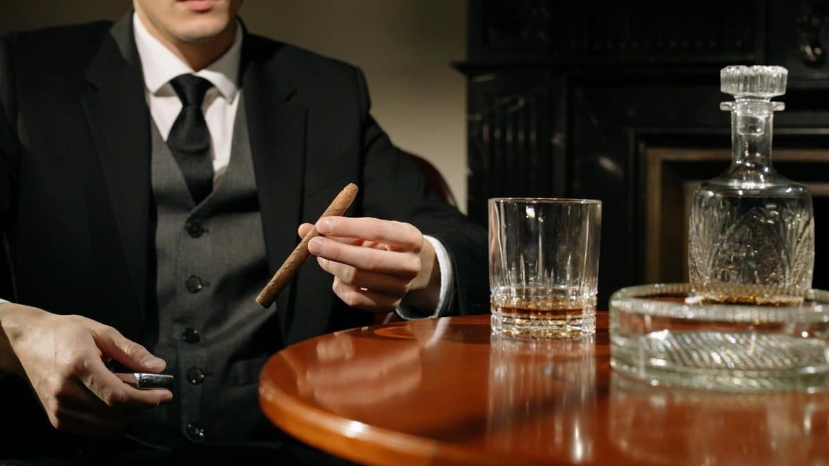 An elegant man in a suit holding a cigar beside a whiskey glass, exuding mystery and class.
