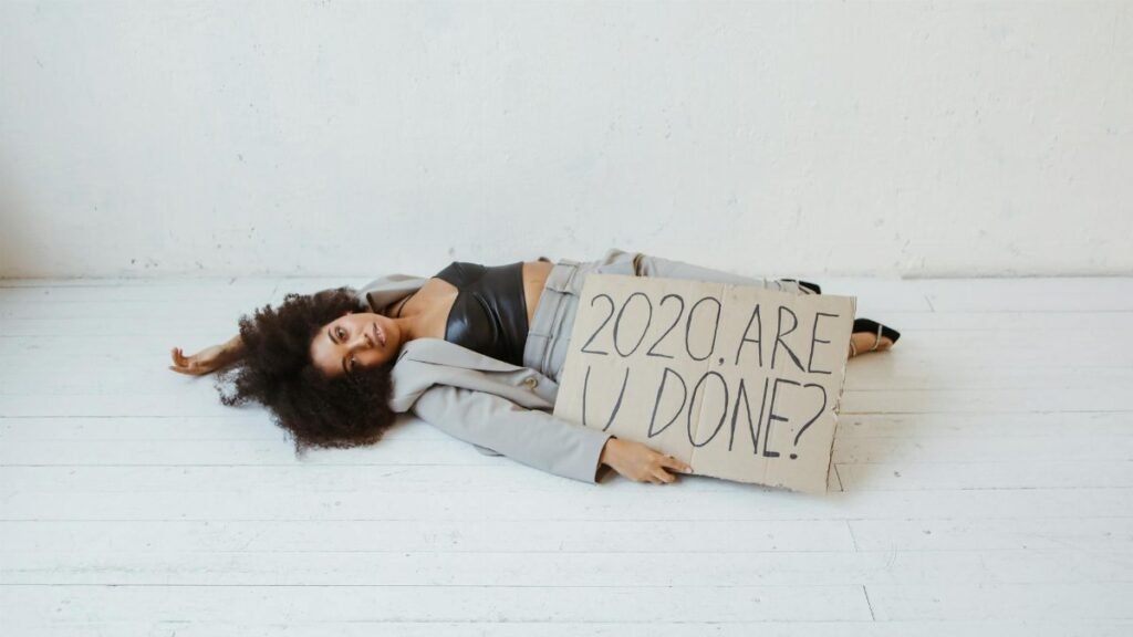 Woman lying on the floor indoors, holding a sign that reads '2020. Are you done?' representing exhaustion and burnout.