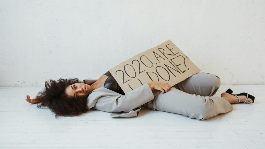 Exhausted woman holding a sign expressing 2020 burnout, lying indoors on a wooden floor.