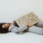 Exhausted woman holding a sign expressing 2020 burnout, lying indoors on a wooden floor.