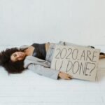 Woman lying on the floor indoors, holding a sign that reads '2020. Are you done?' representing exhaustion and burnout.