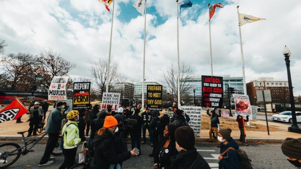 A diverse group of people protests on a Washington DC street with signs and flags on a cloudy day.