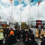 A diverse group of people protests on a Washington DC street with signs and flags on a cloudy day.