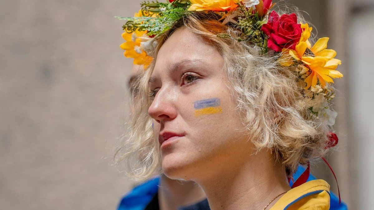 A woman with a flower crown and painted flag on her face shows deep emotion.