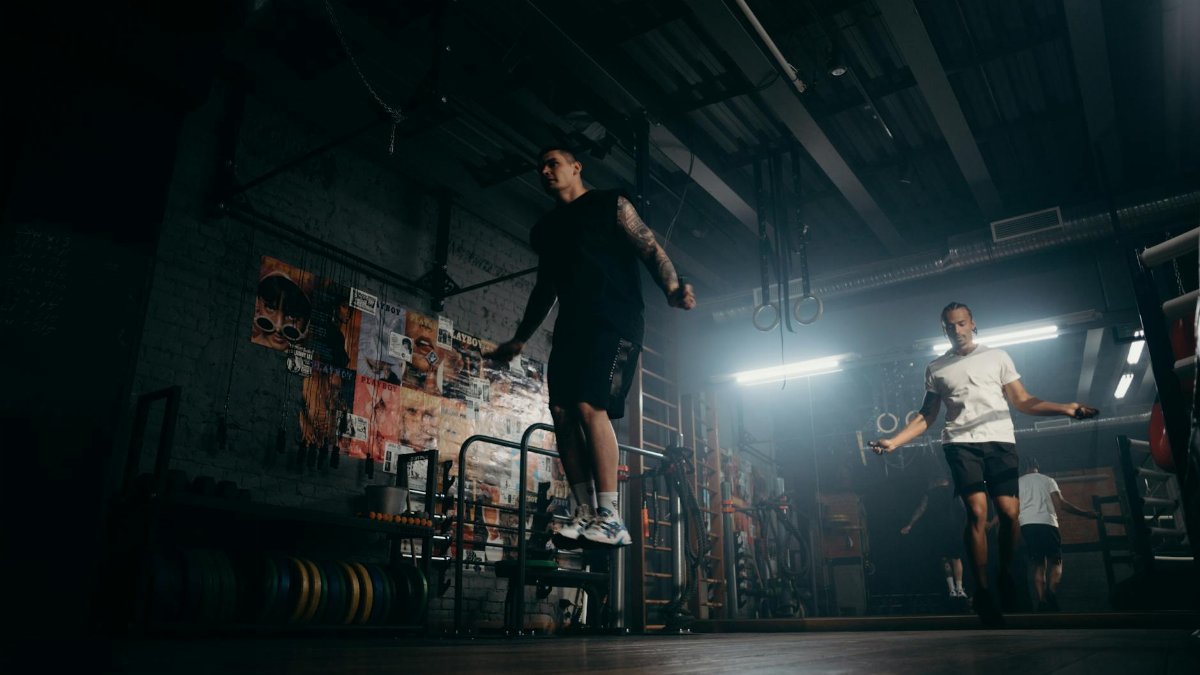 Two men actively skipping jump ropes in a dimly lit gym environment.