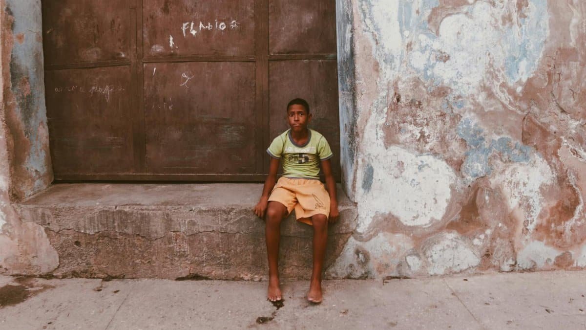 Teen boy sitting on a step in front of a weathered wall in a city street setting.