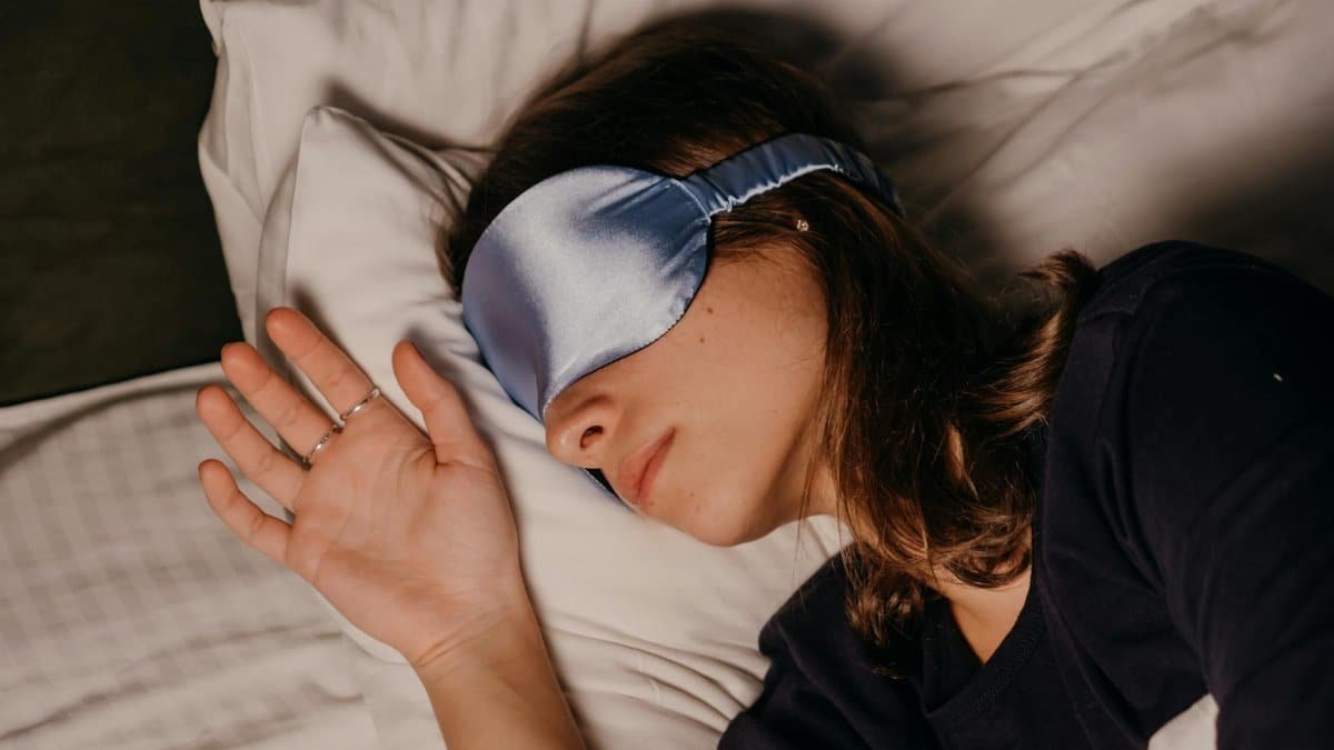 A woman rests peacefully indoors wearing a blue silk sleep mask.