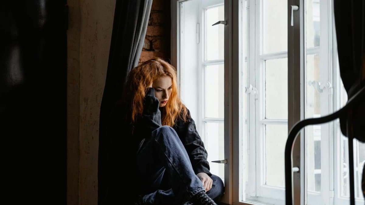 A woman sits by a window, lost in deep thoughts and moody light.