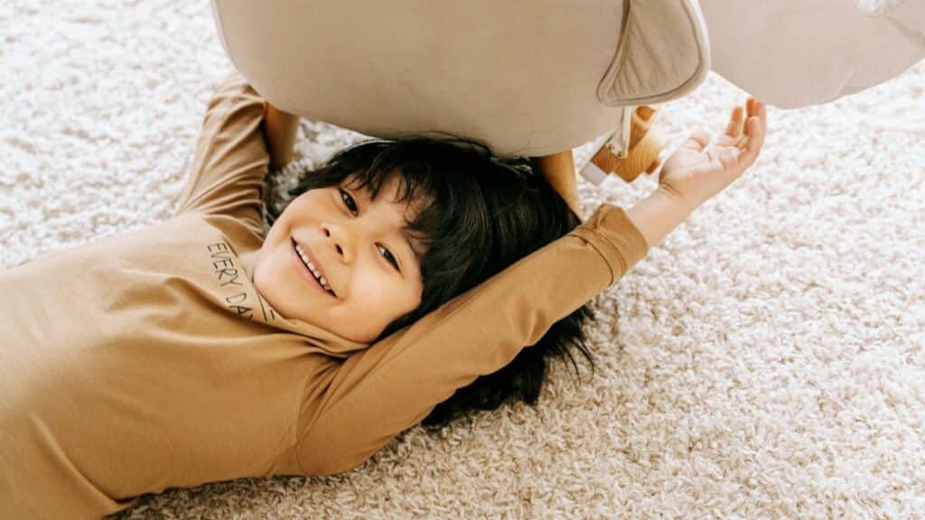 Smiling child lying under a chair on a plush carpet, enjoying a moment of playful relaxation.