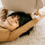 Smiling child lying under a chair on a plush carpet, enjoying a moment of playful relaxation.