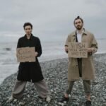 A couple stands on a pebble beach holding signs addressing calmness and longing. Moody atmosphere.