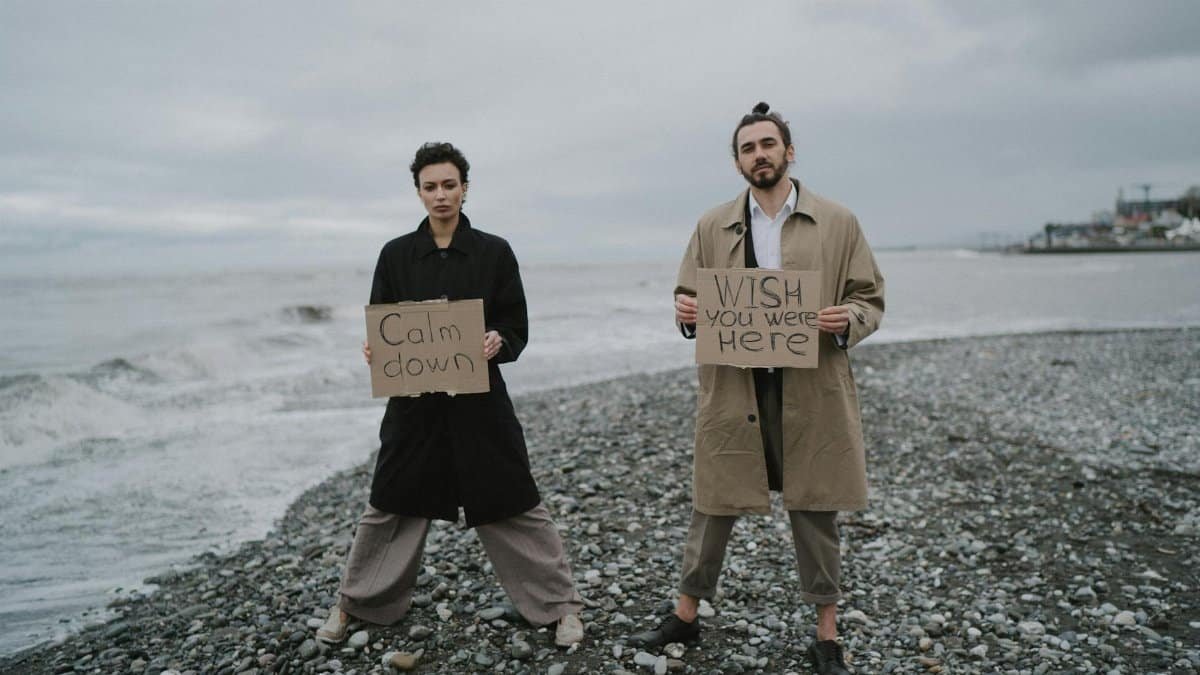 A couple stands on a pebble beach holding signs addressing calmness and longing. Moody atmosphere.