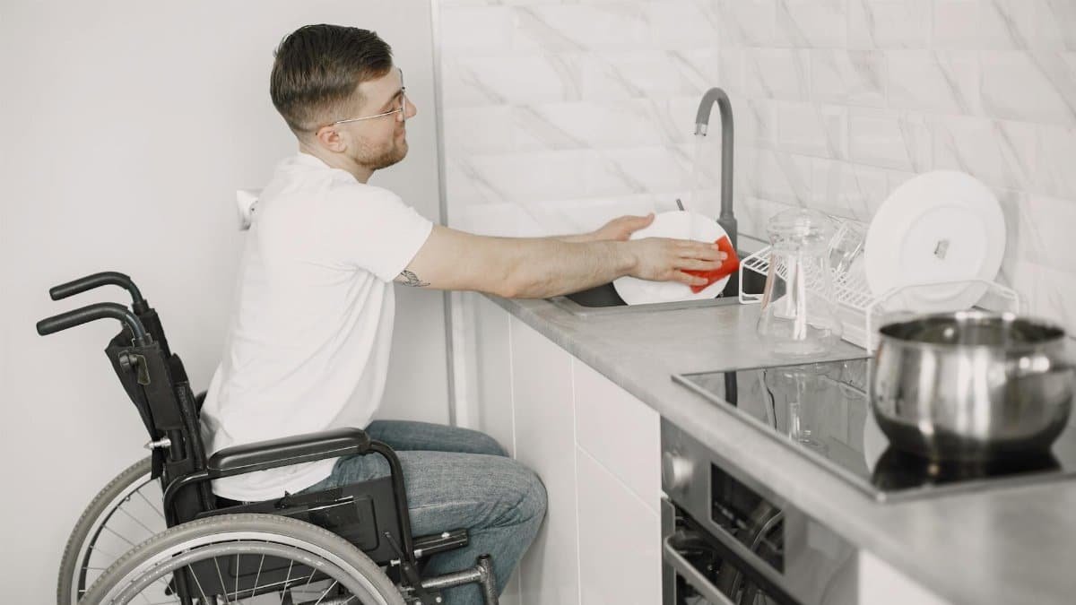 A young man in a wheelchair washing dishes in a modern kitchen, promoting accessibility.