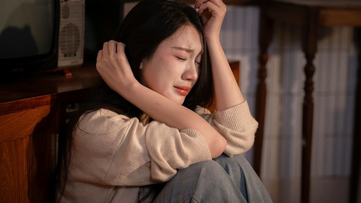 A young woman sitting indoors, expressing deep emotional distress.