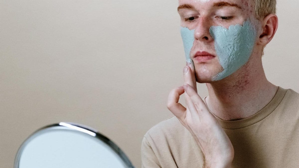 Close-up of a young man applying a clay face mask with a mirror.