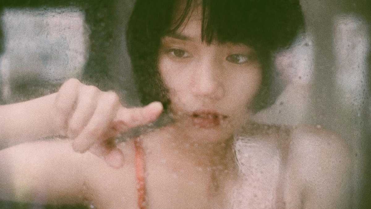 Through window of young Asian woman drawing on wet glass with finger in soft light