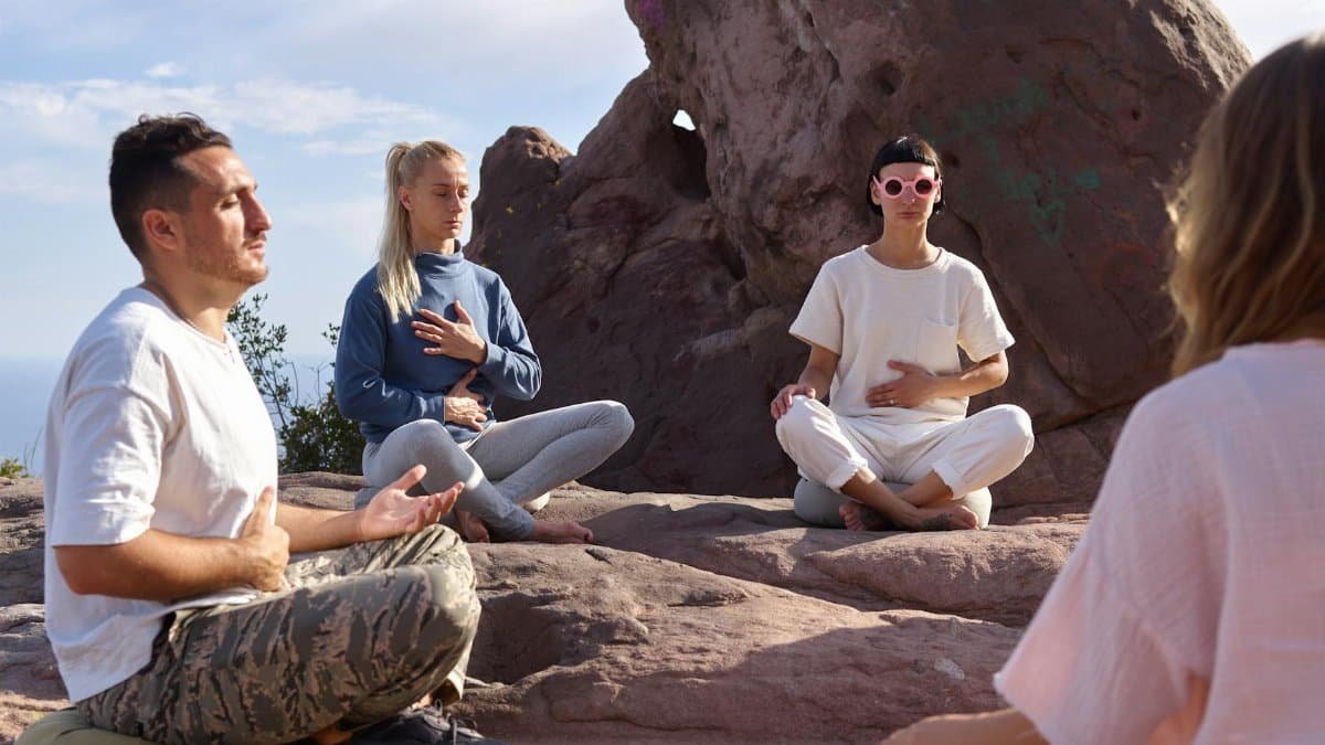 Four people meditate on rocky terrain under a clear sky, promoting serenity and mindfulness.