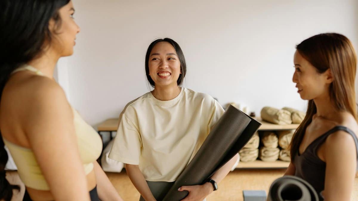 Three women chatting happily indoors, holding yoga mats, embodying friendship and fitness.