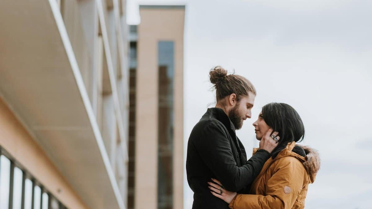 Side view of enamored young bearded man in outerwear touching face of girlfriend and looking at each other near modern building on city street