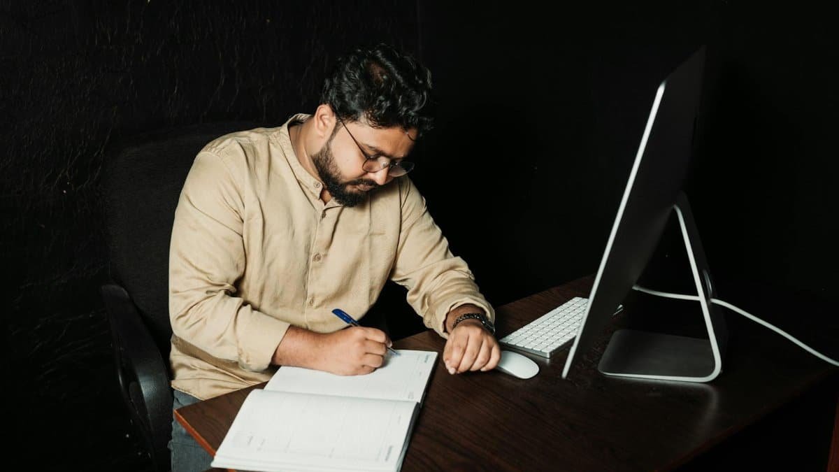 Man writing in office with computer. Late-night work focused on creativity and productivity.