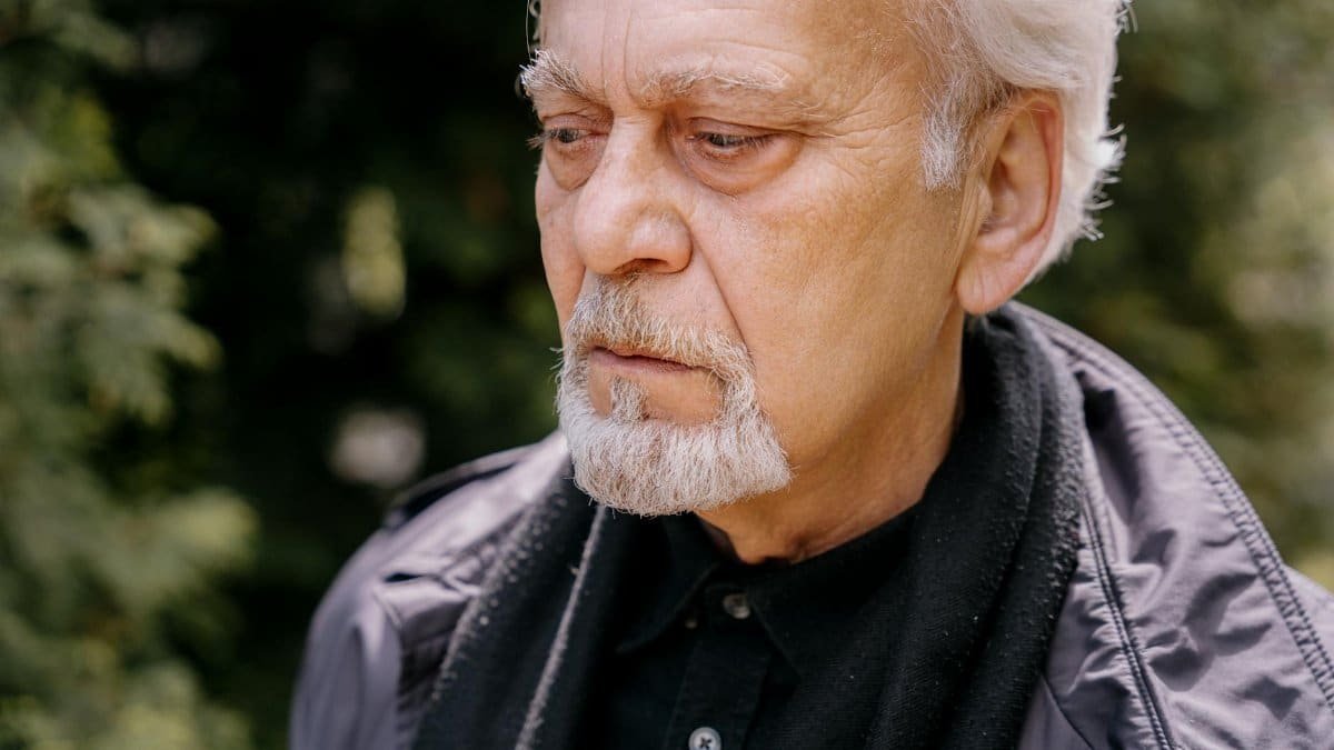 Elderly man with facial hair looking thoughtful outdoors, captured in natural light.