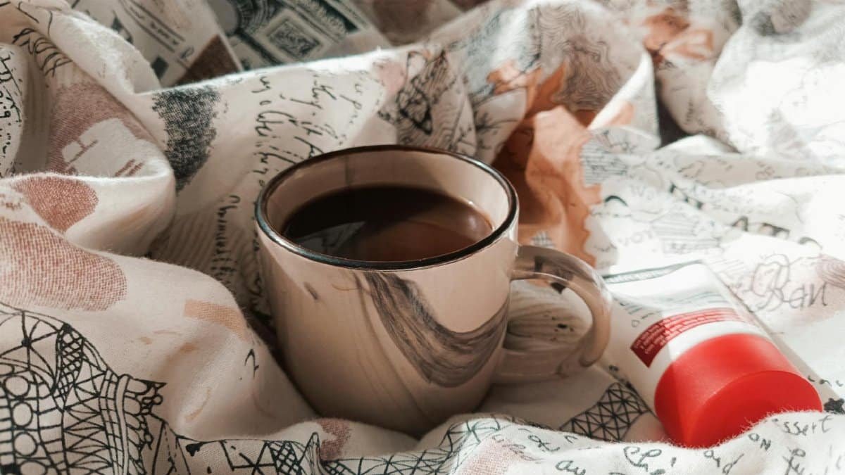 A comfortable setting featuring a steaming cup of coffee and skincare cream on patterned linens.