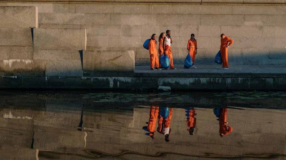 Volunteers in orange uniforms collect trash by the river during an early morning cleanup effort.