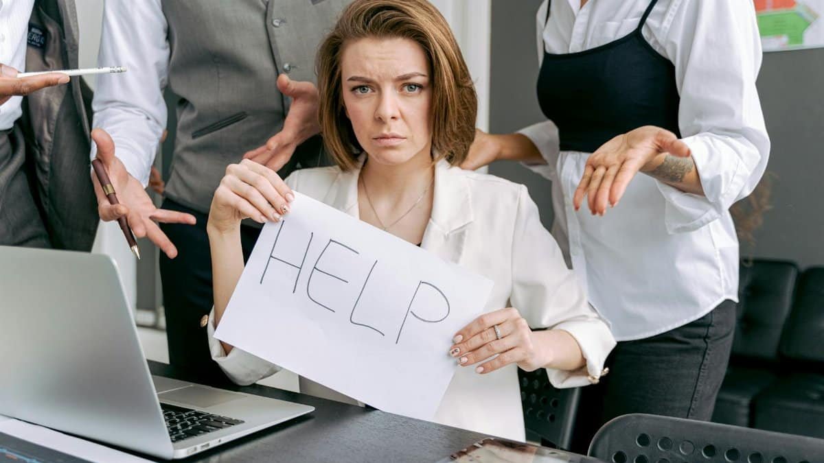 Stressed office worker holding a help sign surrounded by colleagues' demands.