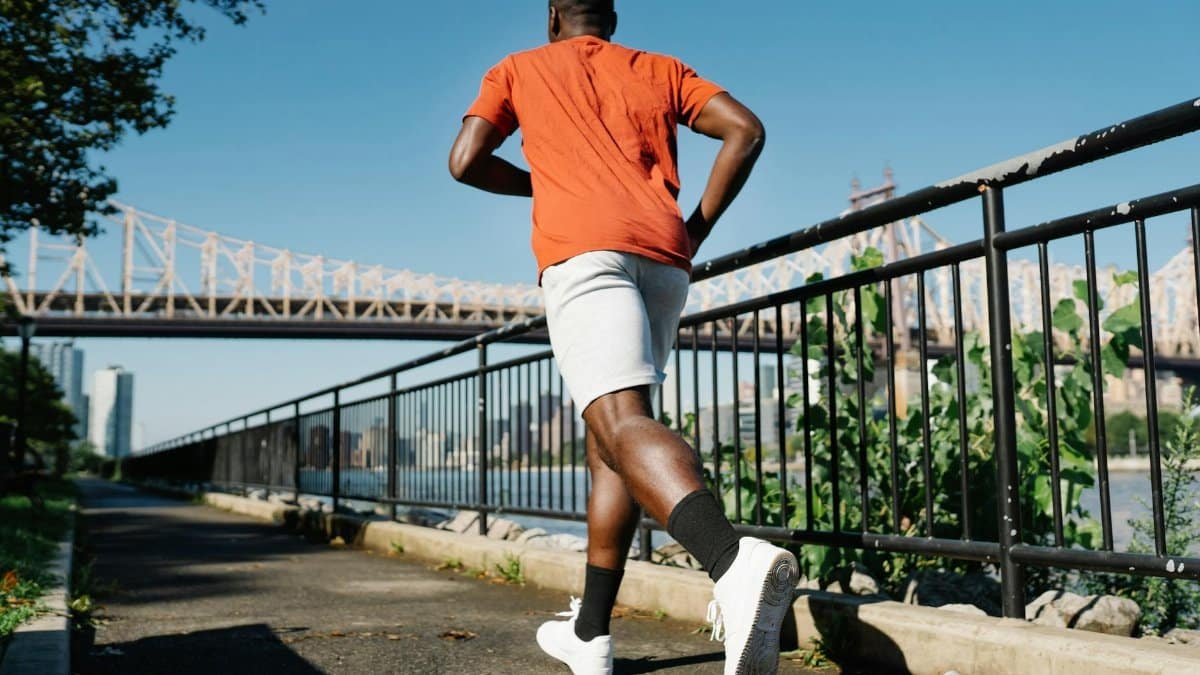 A man jogging on a riverside path, with a city bridge and urban skyline in the background.