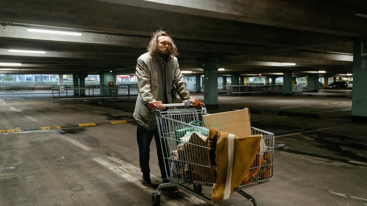 A homeless man pushing a shopping cart filled with belongings in a dimly lit parking garage.