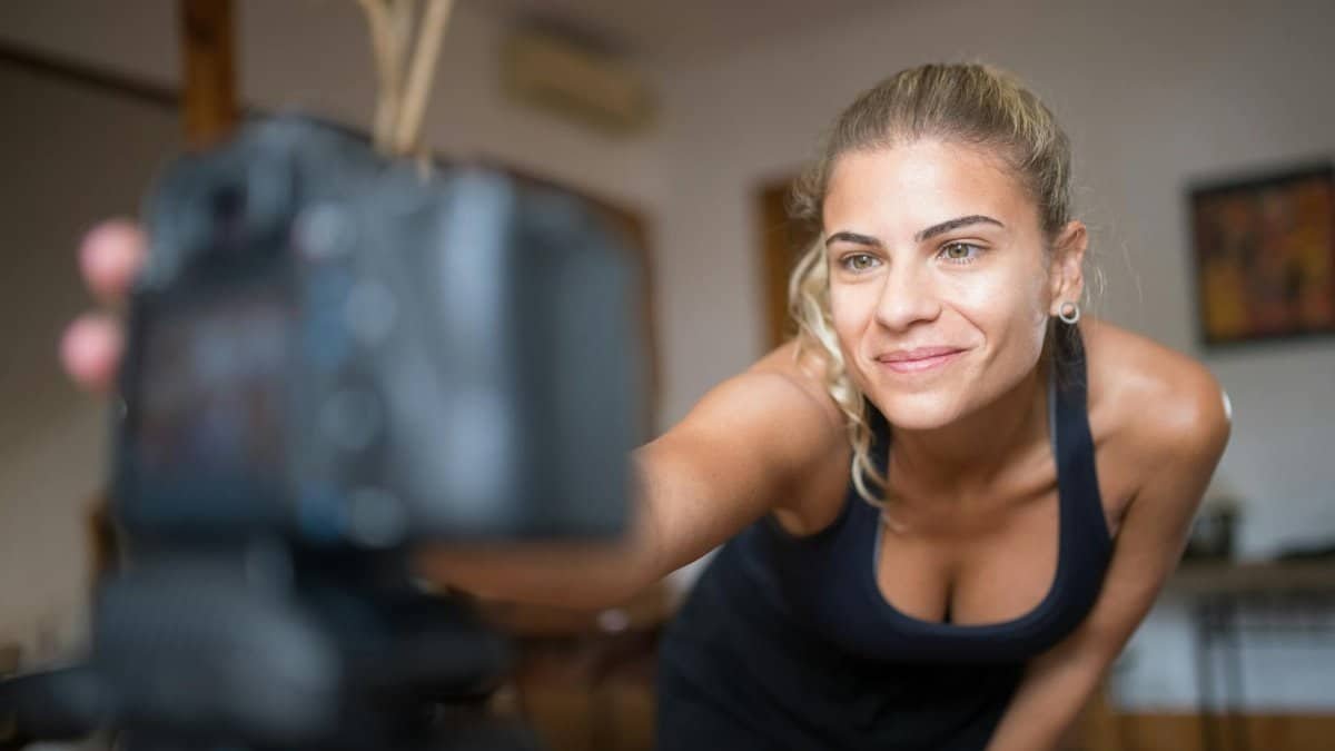 Smiling woman in sportswear setting up a camera indoors for a fitness recording session.