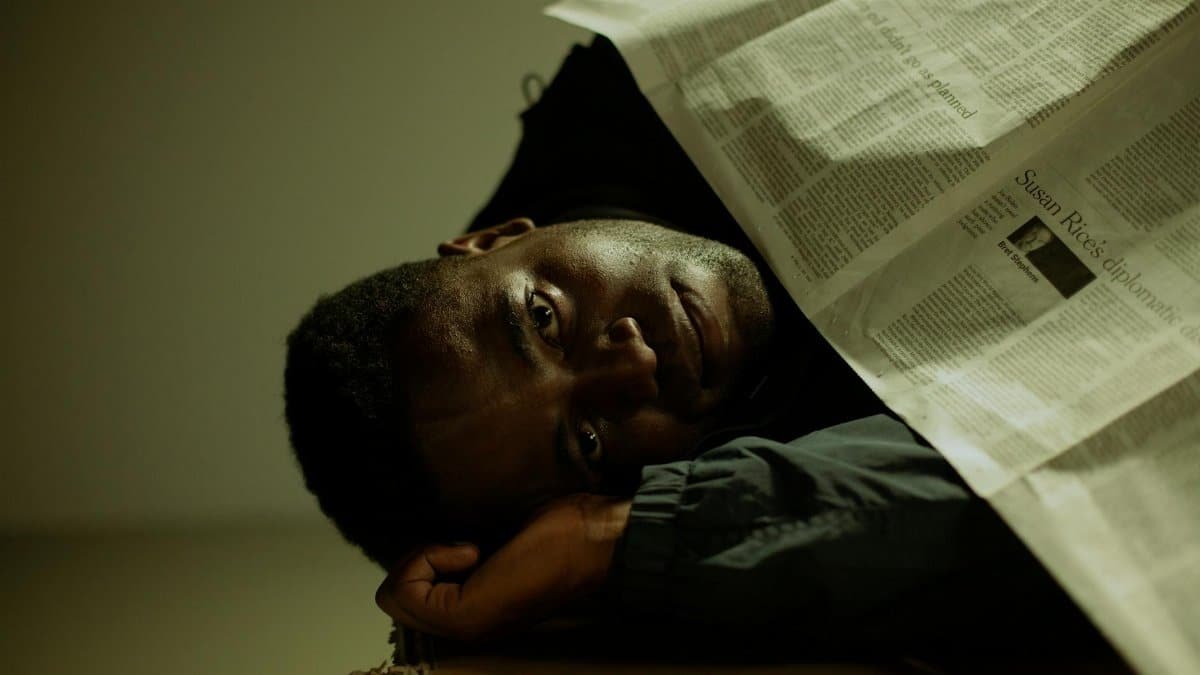 A man rests indoors, covered with a newspaper, symbolizing themes of homelessness and poverty.