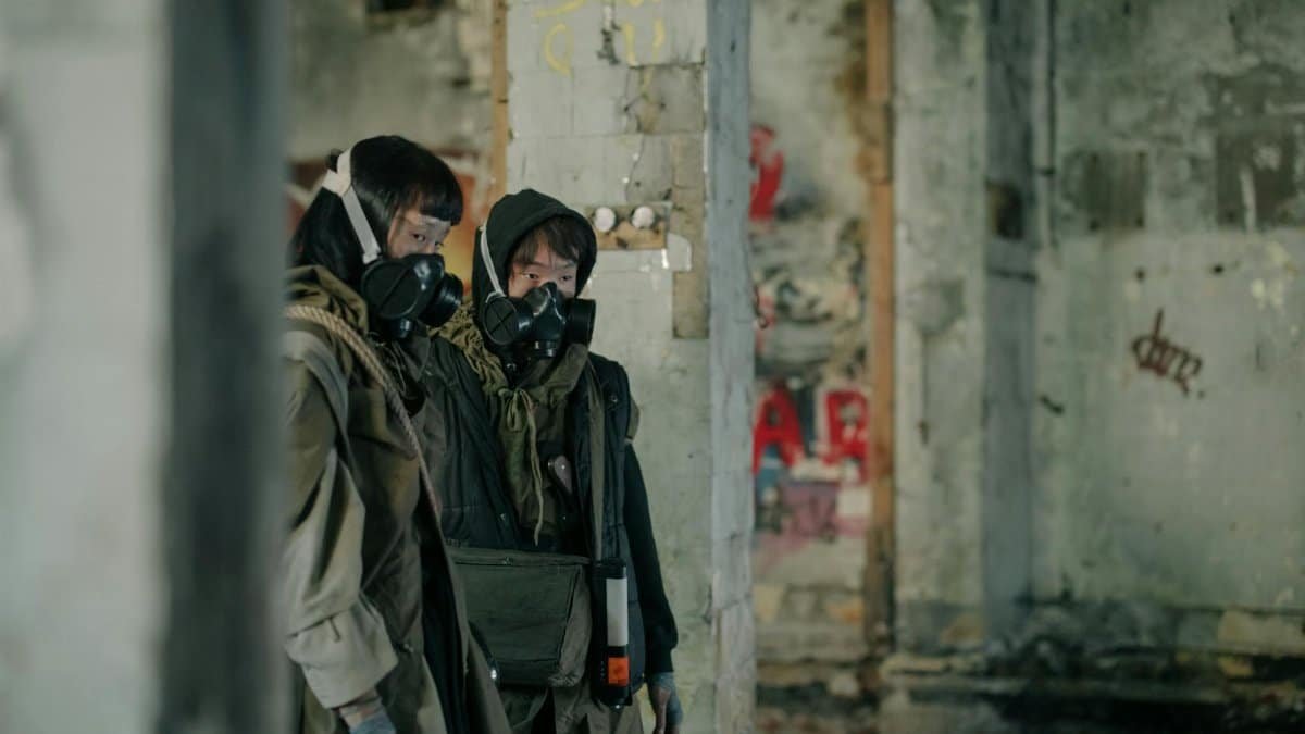 Two people in gas masks explore an abandoned building in a dystopian urban environment.