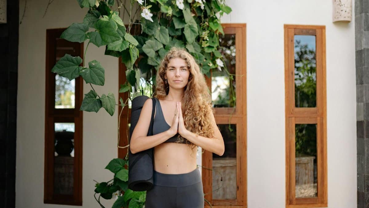 A woman with curly hair standing outdoors in a yoga pose beside a building with ivy.