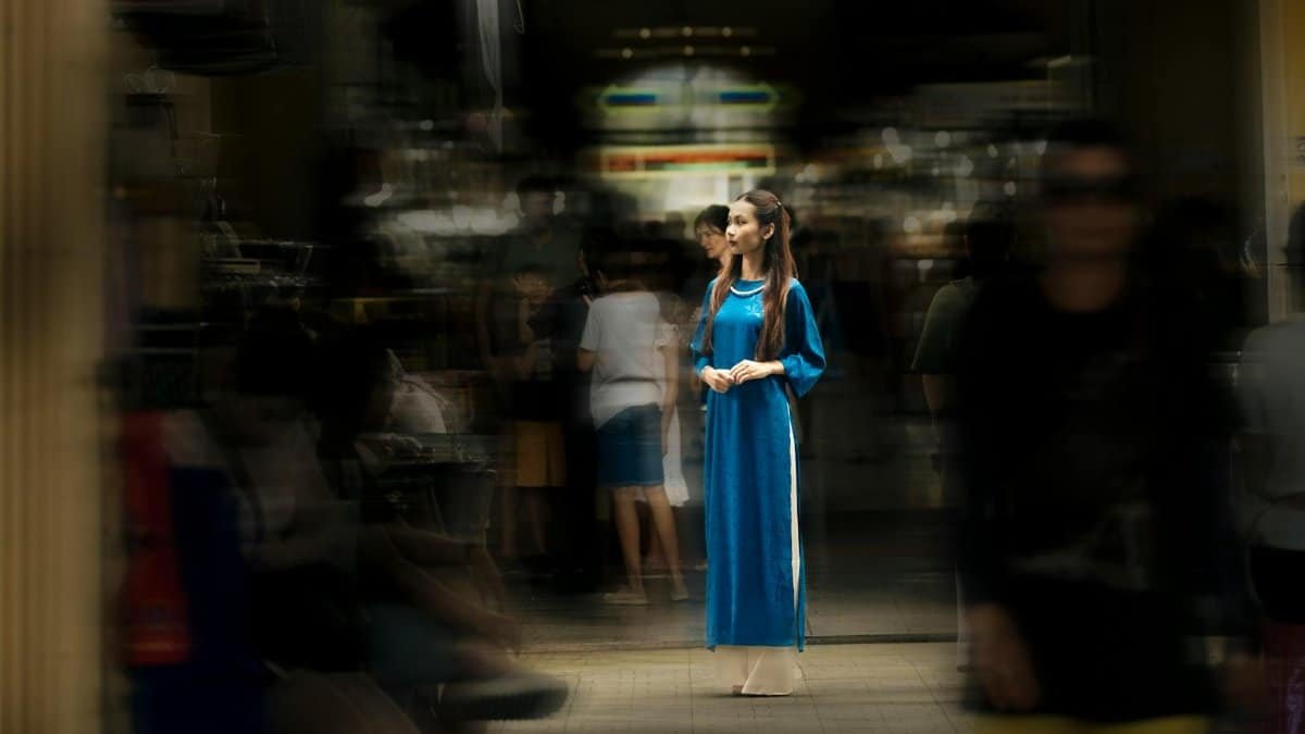 A woman in a blue traditional dress standing still amidst a blurred urban background.