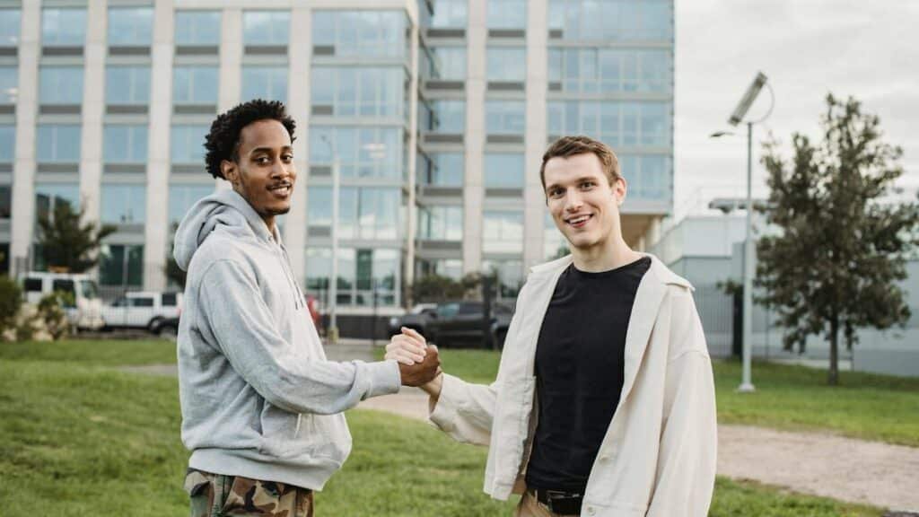 Young diverse male friends shaking hands while standing on lawn against modern building and looking at camera